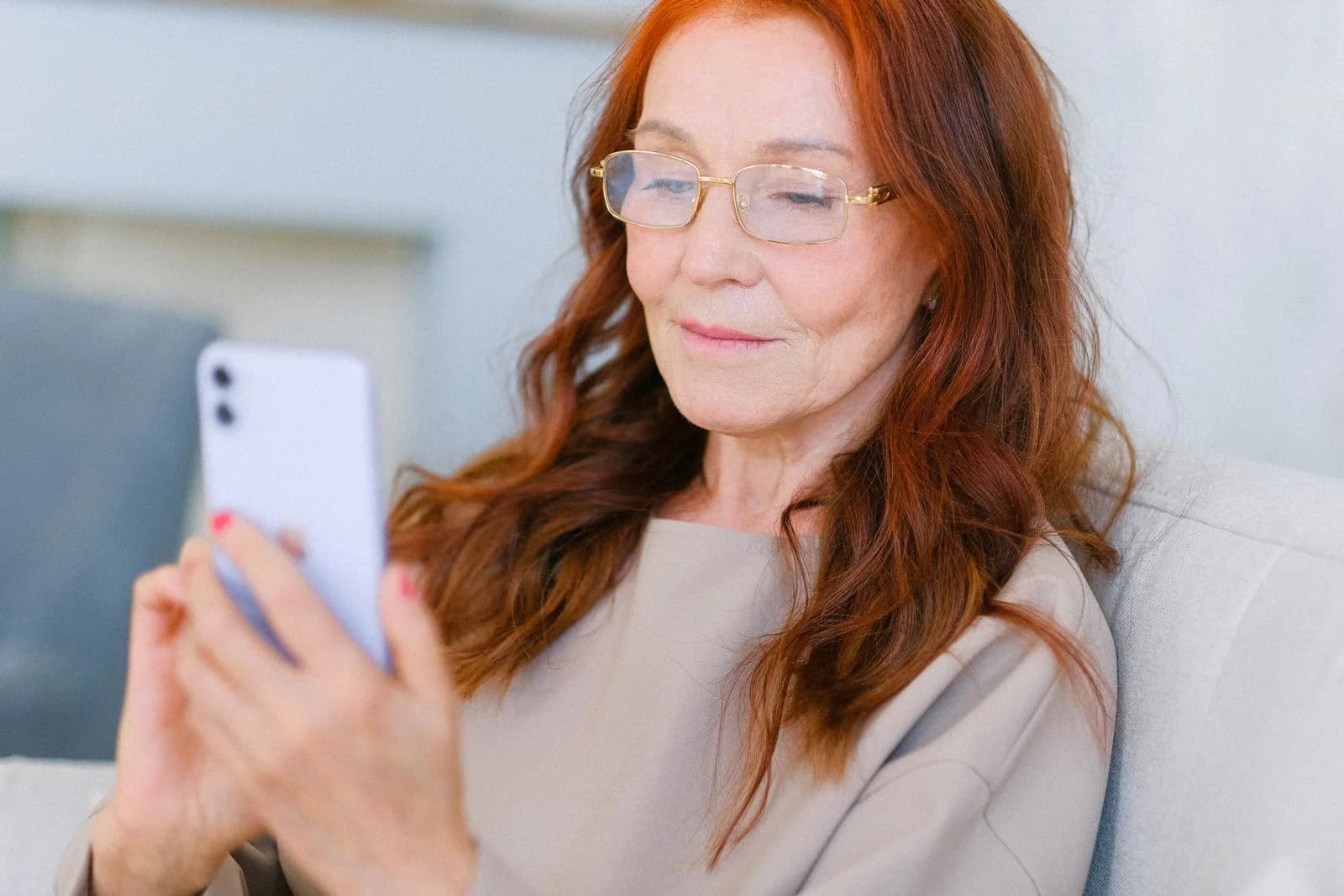 Mulher idosa com cabelo ruivo usando um smartphone e relaxando dentro de casa, ilustrando como cultivar amizades à distância