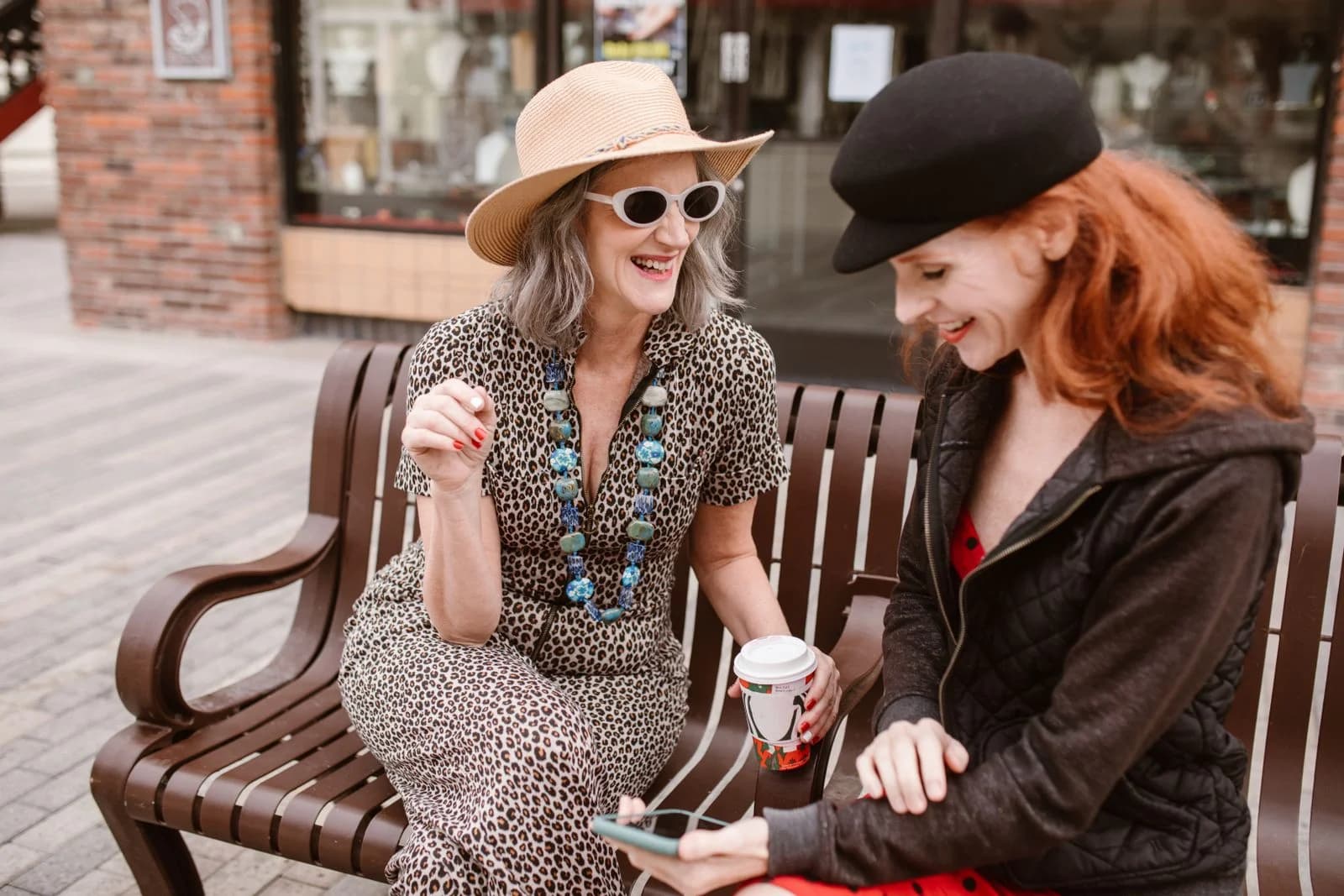 Duas mulheres maduras rindo e tomando café juntas em um banco na rua, ilustrando amizade depois dos 40