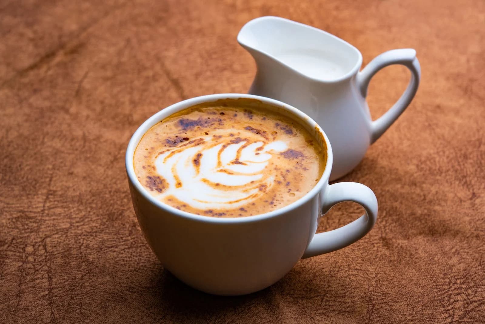 Close-up de cappuccino com arte em latte intricada, acompanhado por uma jarra de creme em uma superfície texturizada, ilustrando gatilhos alimentares que podem piorar os fogachos
