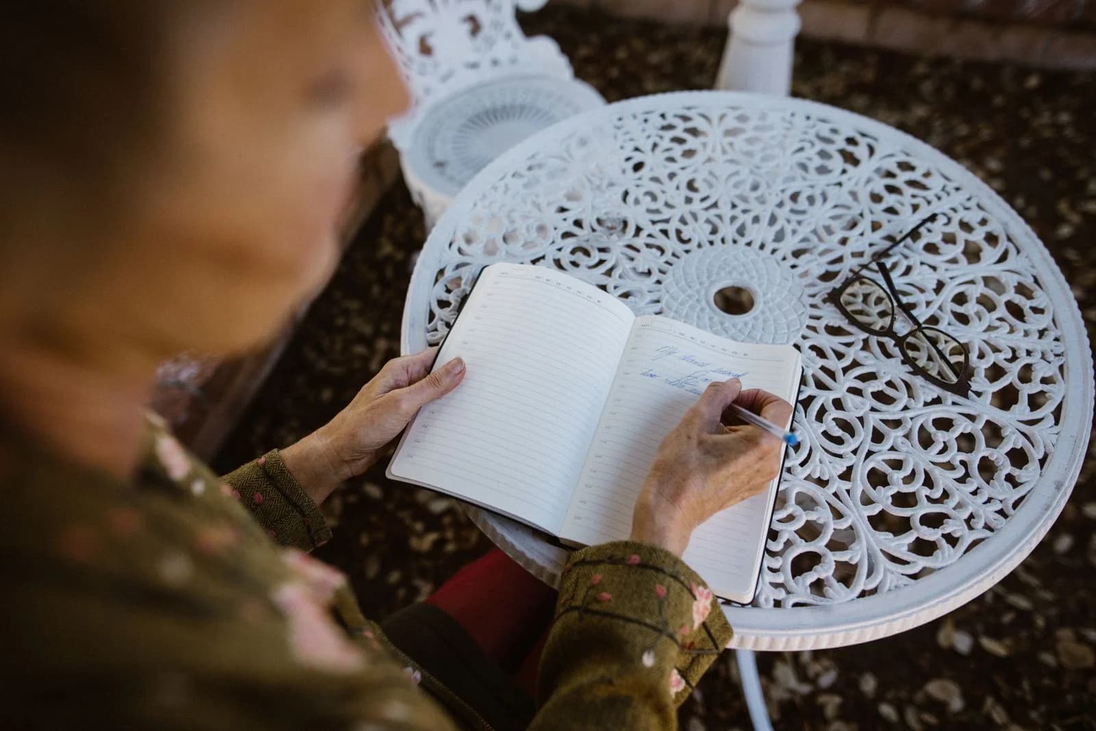 Pessoa idosa escrevendo em um caderno em uma mesa externa ornamentada, capturando um momento pacífico, ilustrando journaling para autoconhecimento