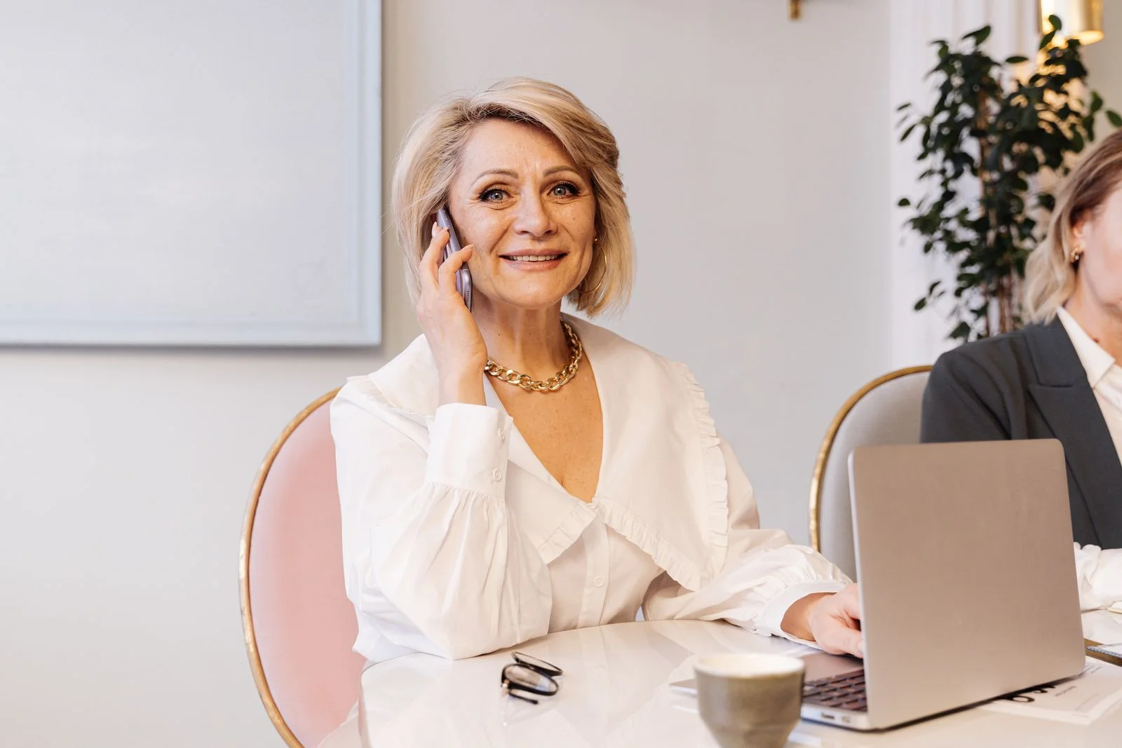 Uma mulher idosa sorridente em uma blusa branca falando ao telefone enquanto trabalha em um ambiente de escritório moderno, ilustrando como dizer não sem culpa