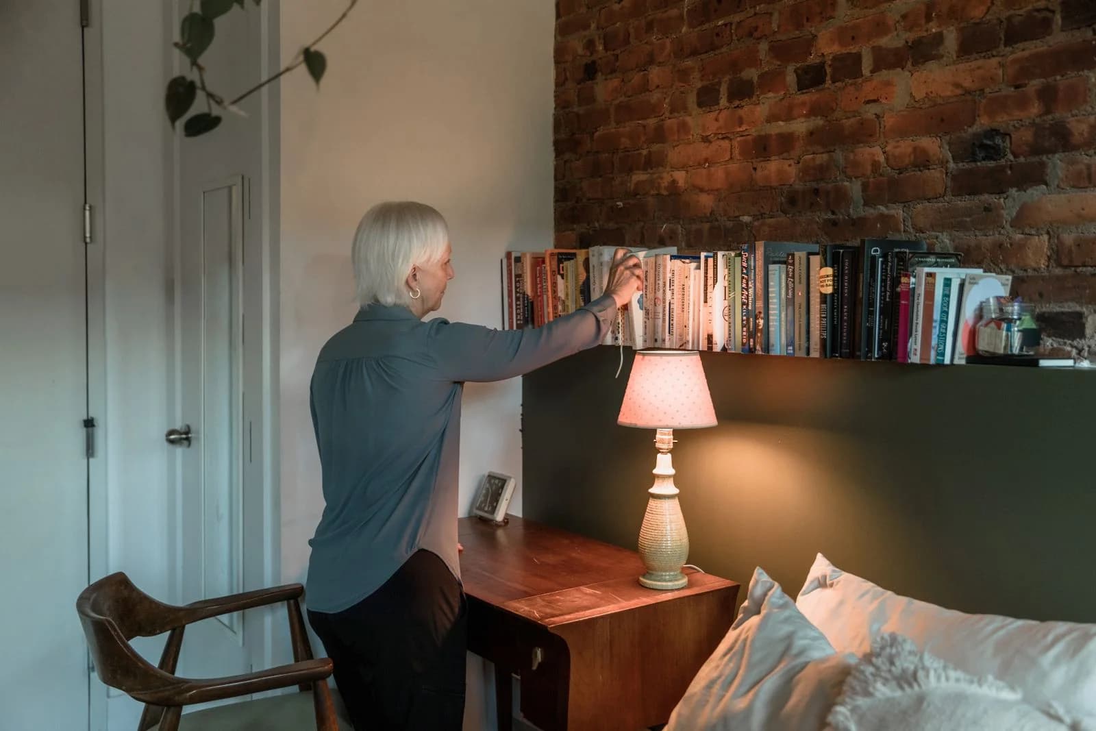 Uma mulher idosa organizando livros em uma estante ao lado de uma lâmpada de mesa em um cômodo quente e aconchegante, ilustrando como reconstruir a rotina diária depois de um divórcio tardio