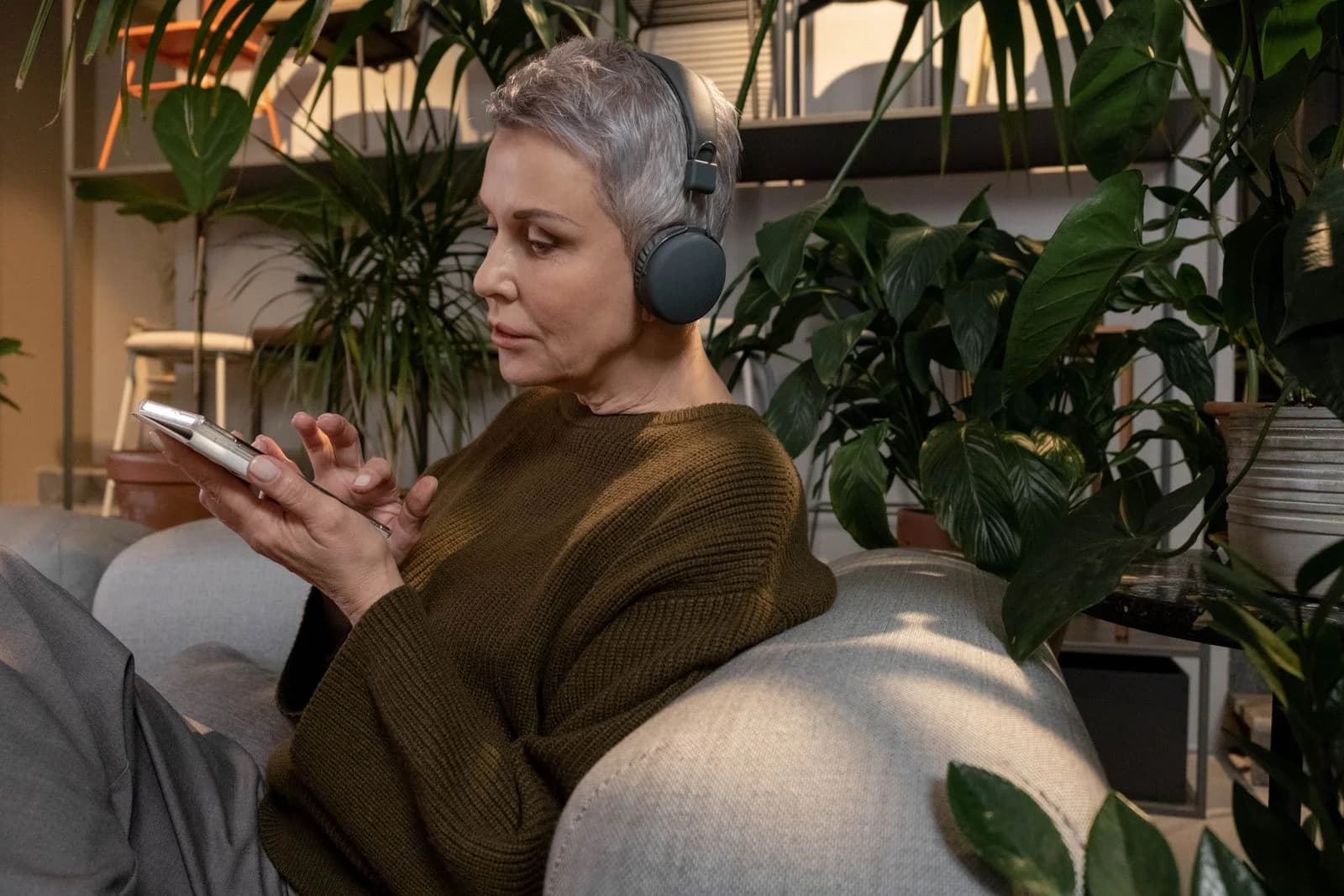 Mulher idosa em uma sala de estar aconchegante usando smartphone com fones de ouvido, cercada por plantas, ilustrando como as redes sociais alimentam a síndrome da impostora