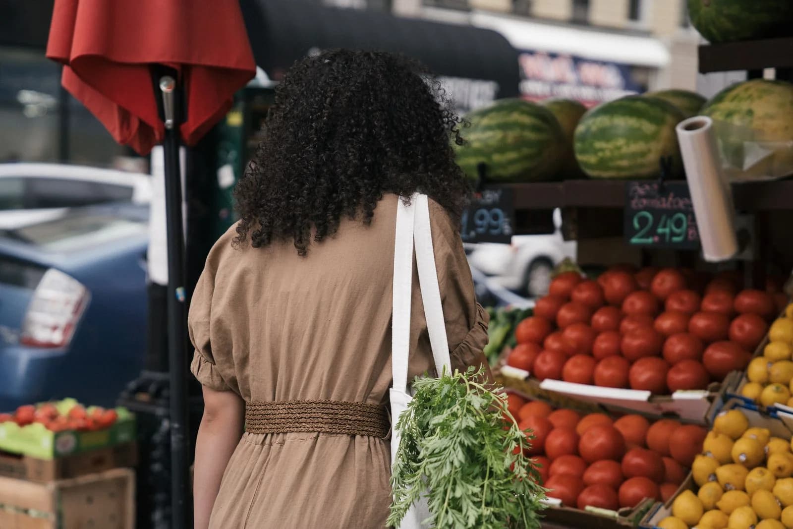 Vista de costas de mulher com bolsa de ombro fazendo compras em um mercado de frutas frescas ao ar livre, ilustrando slow living e consumo consciente