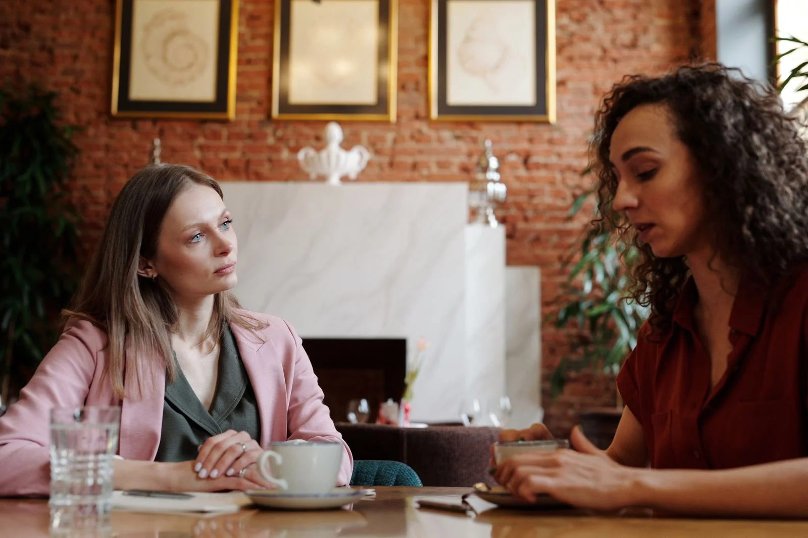 Duas mulheres conversam com seriedade à mesa de um café, tentando resolver um conflito entre amigas