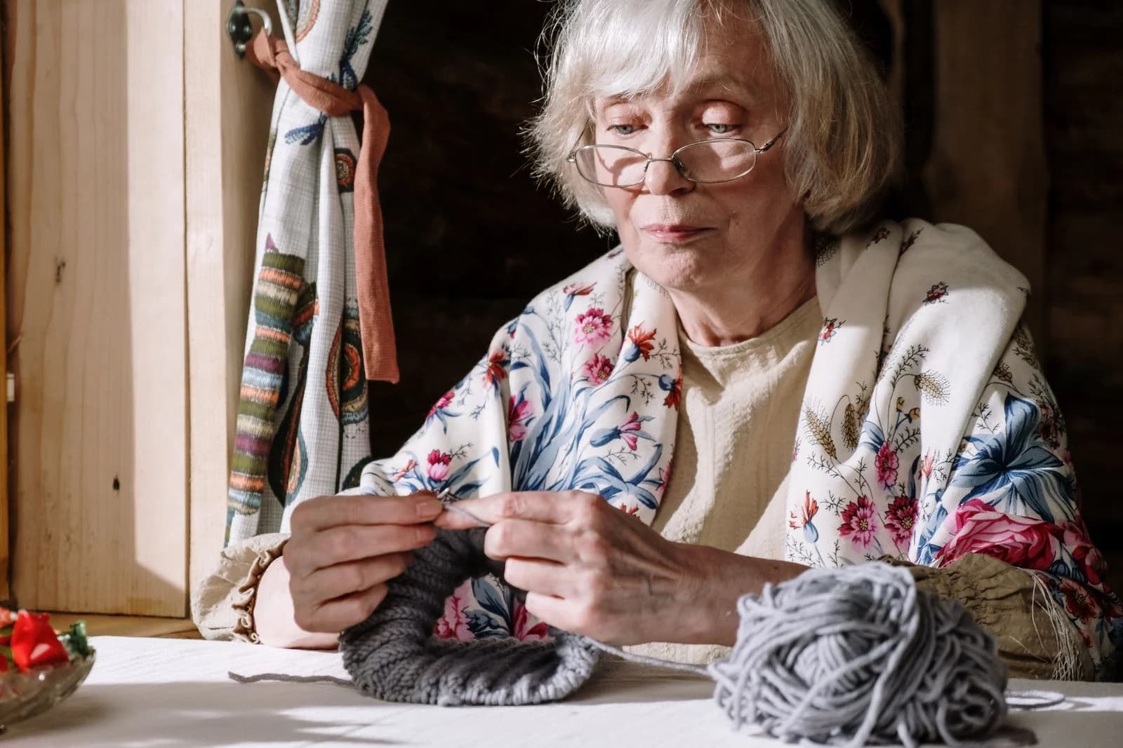 Mulher madura tricota com lã cinza perto da janela, um dos hobbies manuais que acalmam a mente