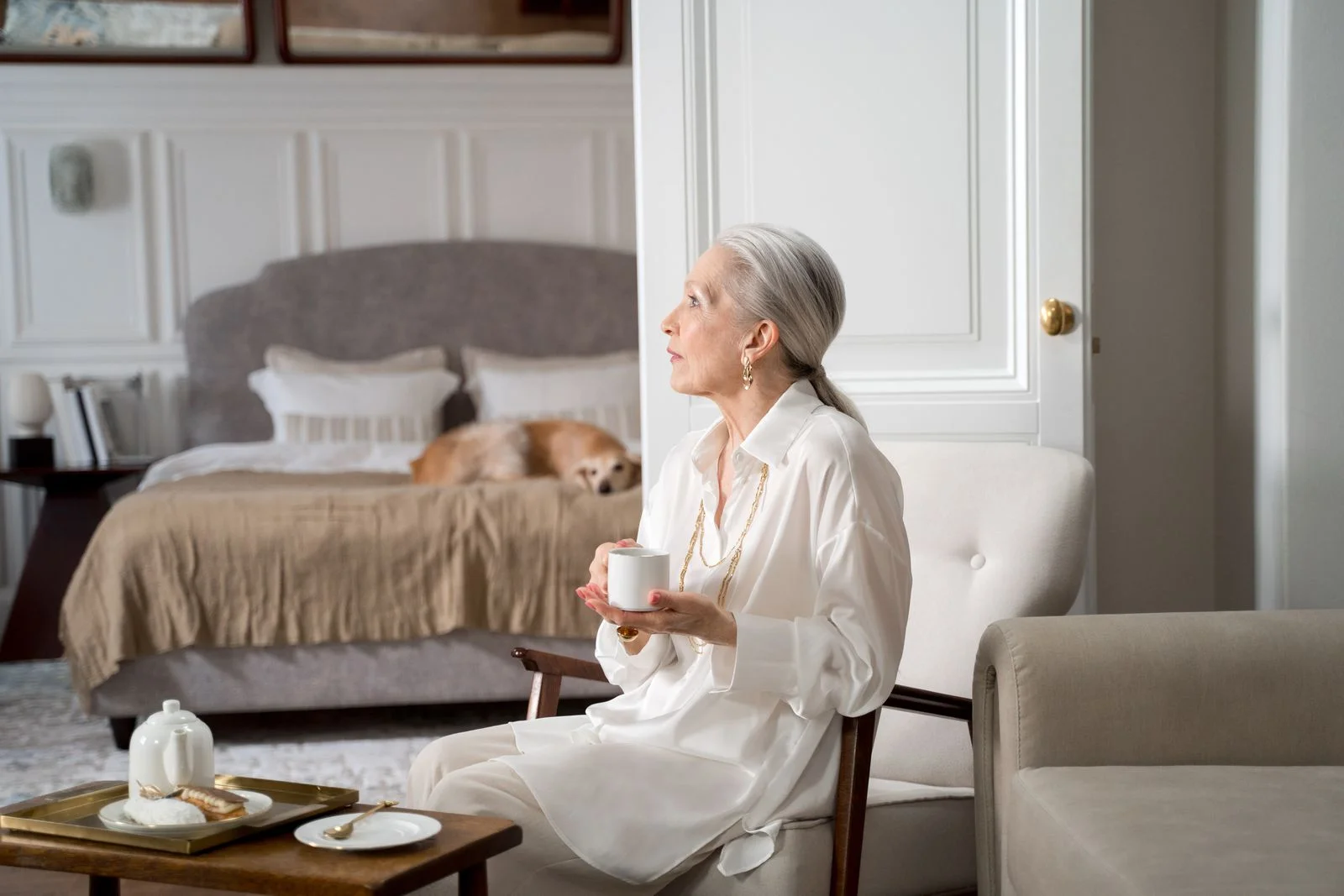Mulher idosa com cabelo grisalho saboreando café em uma sala de estar acolhedora com um cachorro descansando na cama, ilustrando pequenos rituais de slow living no dia a dia