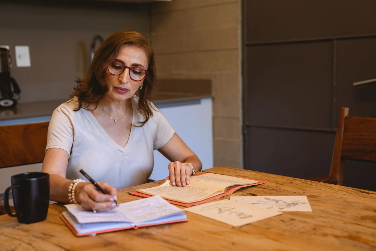 Mulher madura de óculos escreve em um caderno à mesa da cozinha, refletindo sobre seus valores pessoais depois dos 40